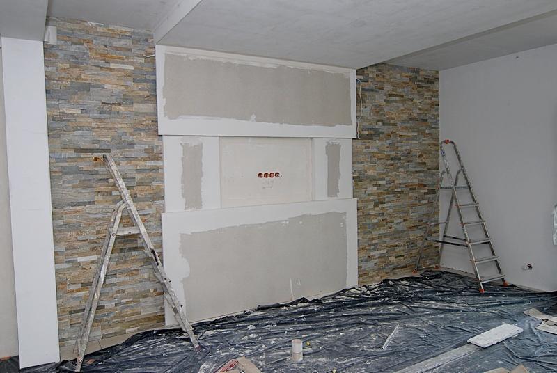 Wnętrze w trakcie prac: ściana z kamienia dekoracyjnego, płyty gipsowe na ścianie, zabezpieczona podłoga folią, dwie drabiny. Widok na etap wykończenia.