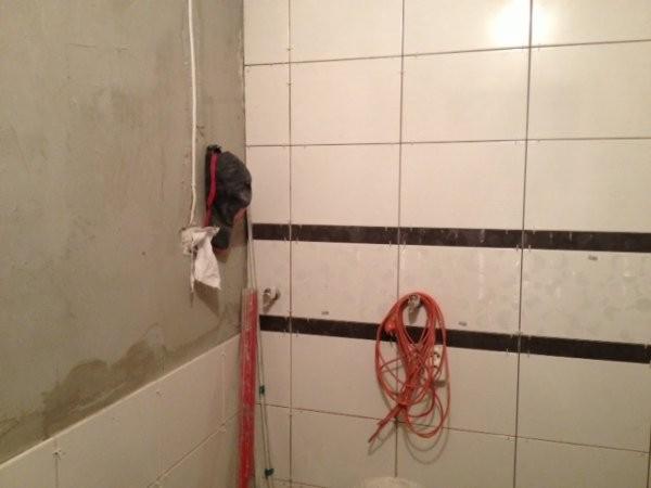 Niedokończona łazienka: białe płytki z czarnym pasem, surowa ściana, kabel i poziomica. Etap prac wykończeniowych, widoczne narzędzia i materiały budowlane.