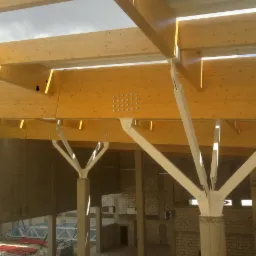 Wnętrze w budowie: drewniane belki stropowe z metalowymi łącznikami, betonowe słupy. Widoczny fragment ściany z cegły i konstrukcja dachu. Jasne, naturalne światło.