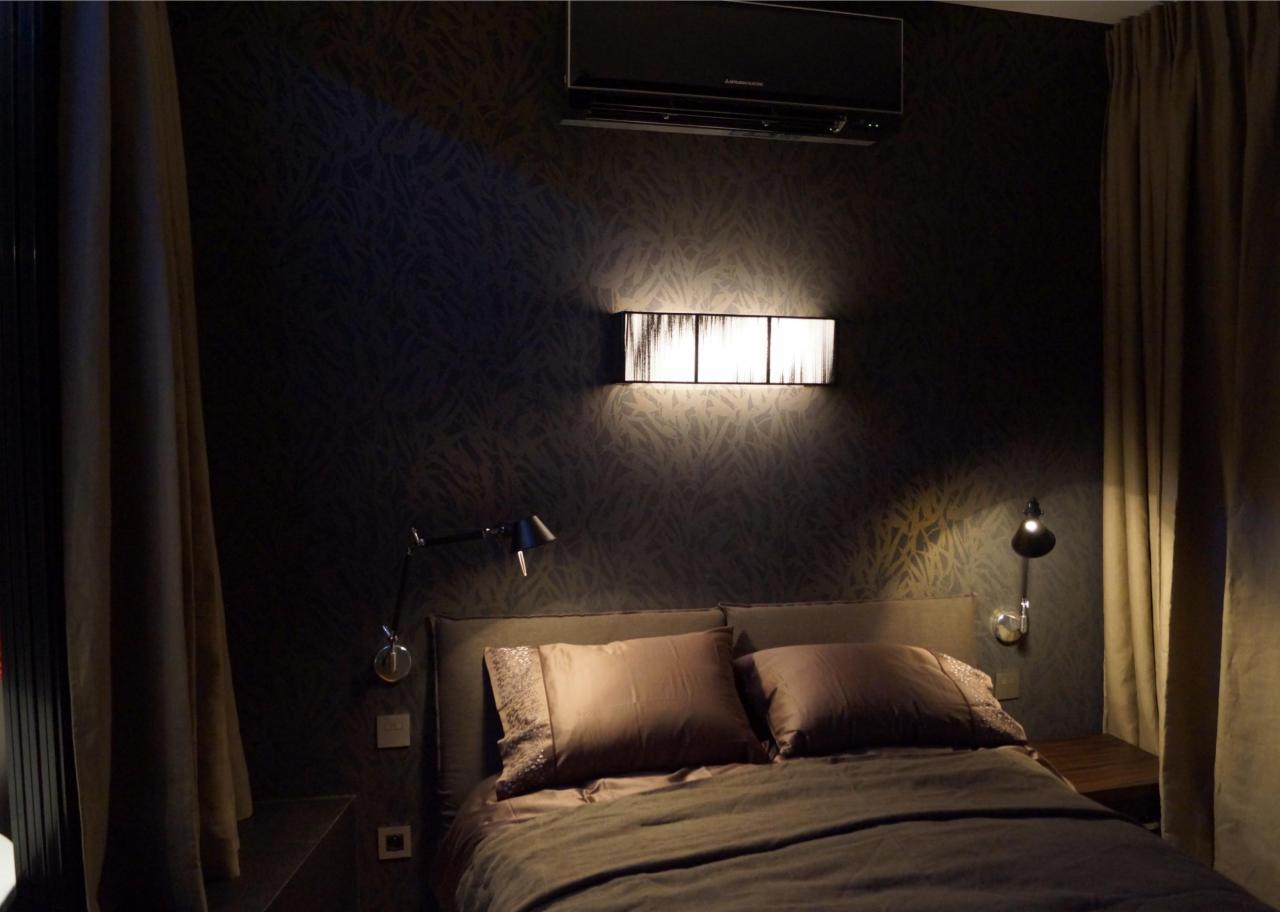Sypialnia w ciemnej tonacji z wzorzystą tapetą, kinkietem nad łóżkiem, lampką nocną na elastycznym ramieniu i klimatyzatorem.