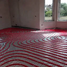 Czerwone rurki ogrzewania podłogowego ułożone na folii i siatce w surowym pomieszczeniu z oknami.