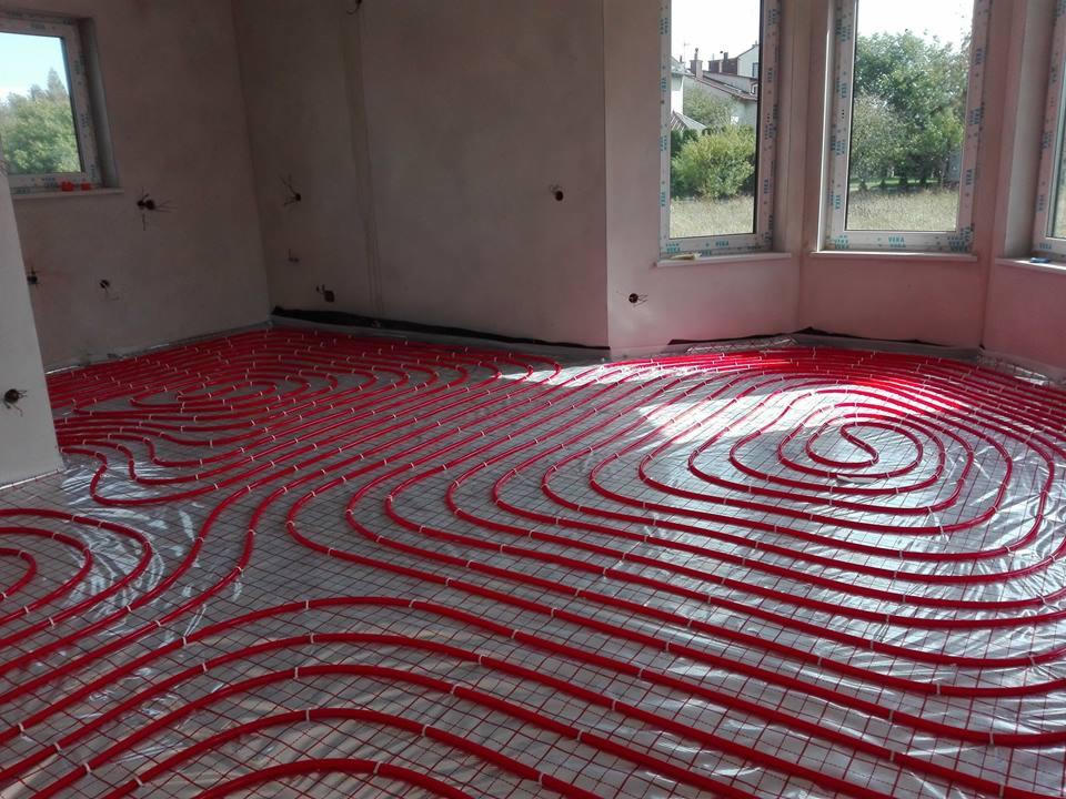 Czerwone rurki ogrzewania podłogowego ułożone na folii i siatce w surowym pomieszczeniu z oknami.