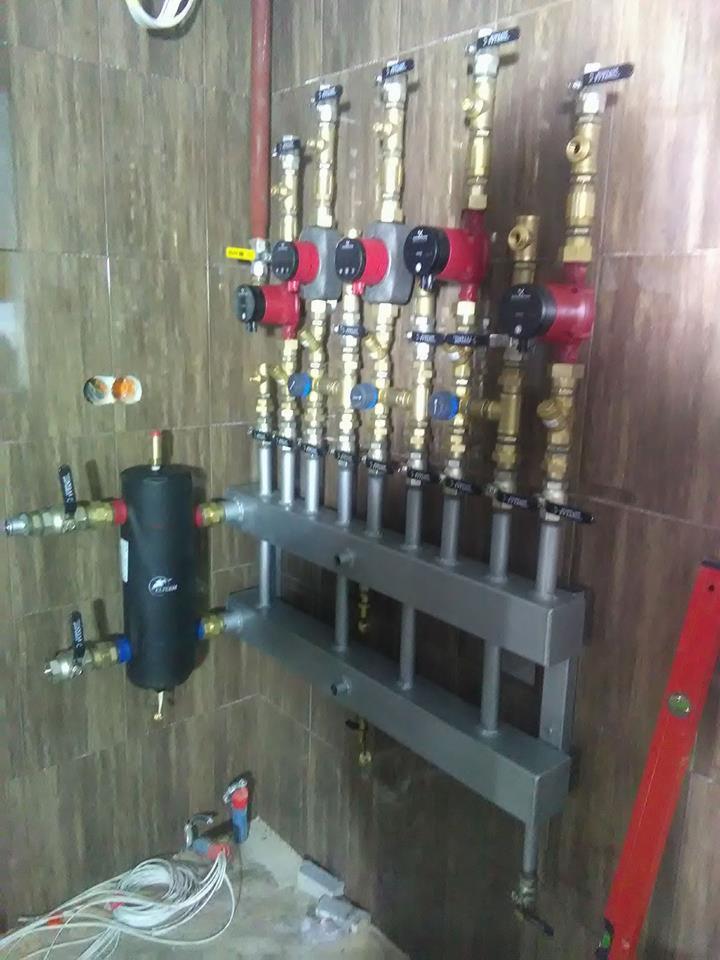 Rozdzielacz ogrzewania podłogowego z pompami obiegowymi, zaworami i zbiornikiem wyrównawczym zamontowany na ścianie z brązowych płytek. Widoczne przewody elektryczne i poziomica.