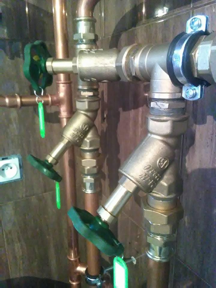 Miedziane i metalowe rury z zielonymi zaworami, połączone z instalacją wodną w pomieszczeniu z ciemnymi płytkami.