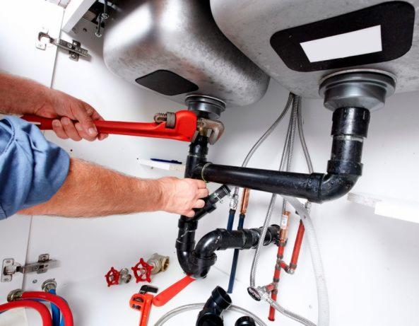 Hydraulik dokręca czerwoną żabką czarne rury pod zlewem w szafce, widoczne liczne przewody i zawory.