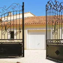 Otwarta, kuta brama wjazdowa z dekoracyjnymi elementami na tle domu z garażem i błękitnym niebem. Widoczny fragment muru z lewej strony.