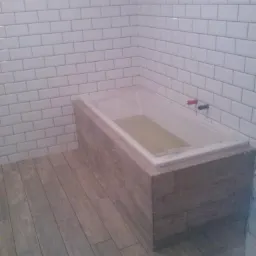 Wanna obudowana płytkami imitującymi drewno, białe płytki na ścianie, podłoga wyłożona płytkami w odcieniach szarości, widoczne niedokończone prace na górze ściany.