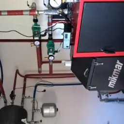 Czarno-czerwony piec z zainstalowaną instalacją grzewczą, widoczne rury w izolacji, pompy obiegowe, manometry i zbiorniki wyrównawcze.