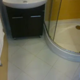 Jasna łazienka z kabiną prysznicową i umywalką z szafką. Białe płytki podłogowe ułożone pod kątem. Żółte płytki ścienne w kabinie. Umywalka z baterią.