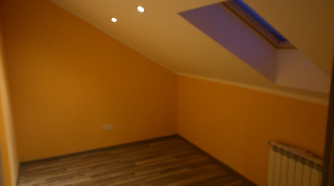 Poddasze po malowaniu: pomarańczowe ściany, drewnopodobna podłoga, okno dachowe i grzejnik. Widoczne trzy halogeny w skosie sufitu.