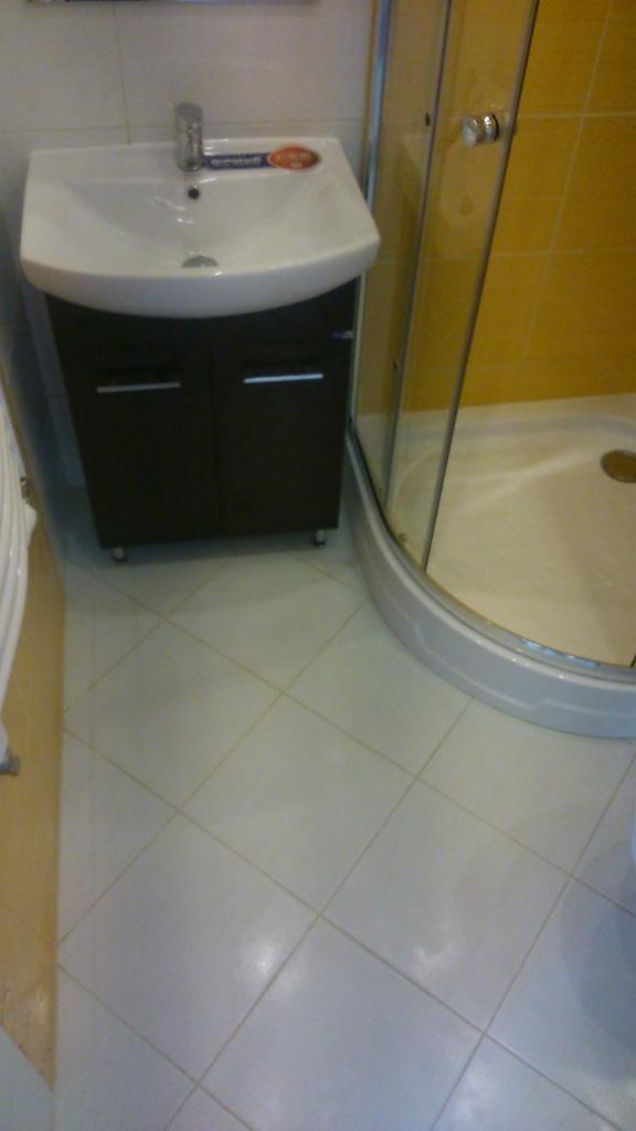 Jasna łazienka z kabiną prysznicową i umywalką z szafką. Białe płytki podłogowe ułożone pod kątem. Żółte płytki ścienne w kabinie. Umywalka z baterią.