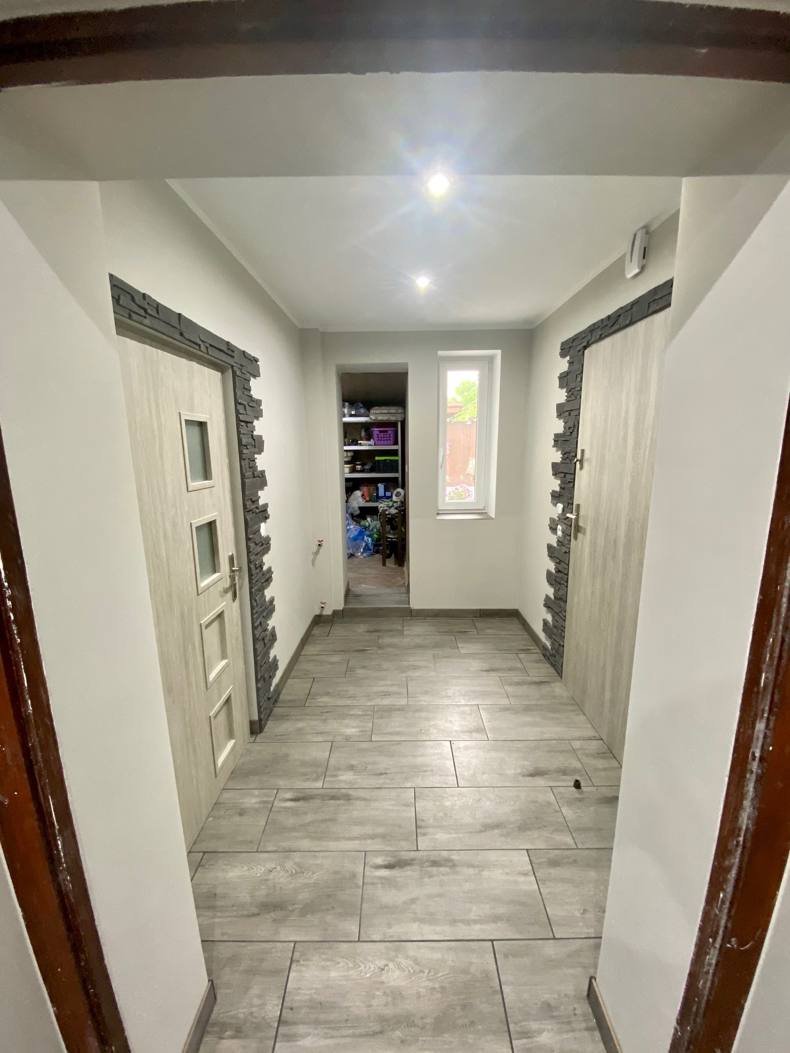 Wykończony korytarz z szarymi płytkami podłogowymi imitującymi drewno, drzwi z kamienną okładziną i widokiem na zagraconą spiżarnię w głębi.