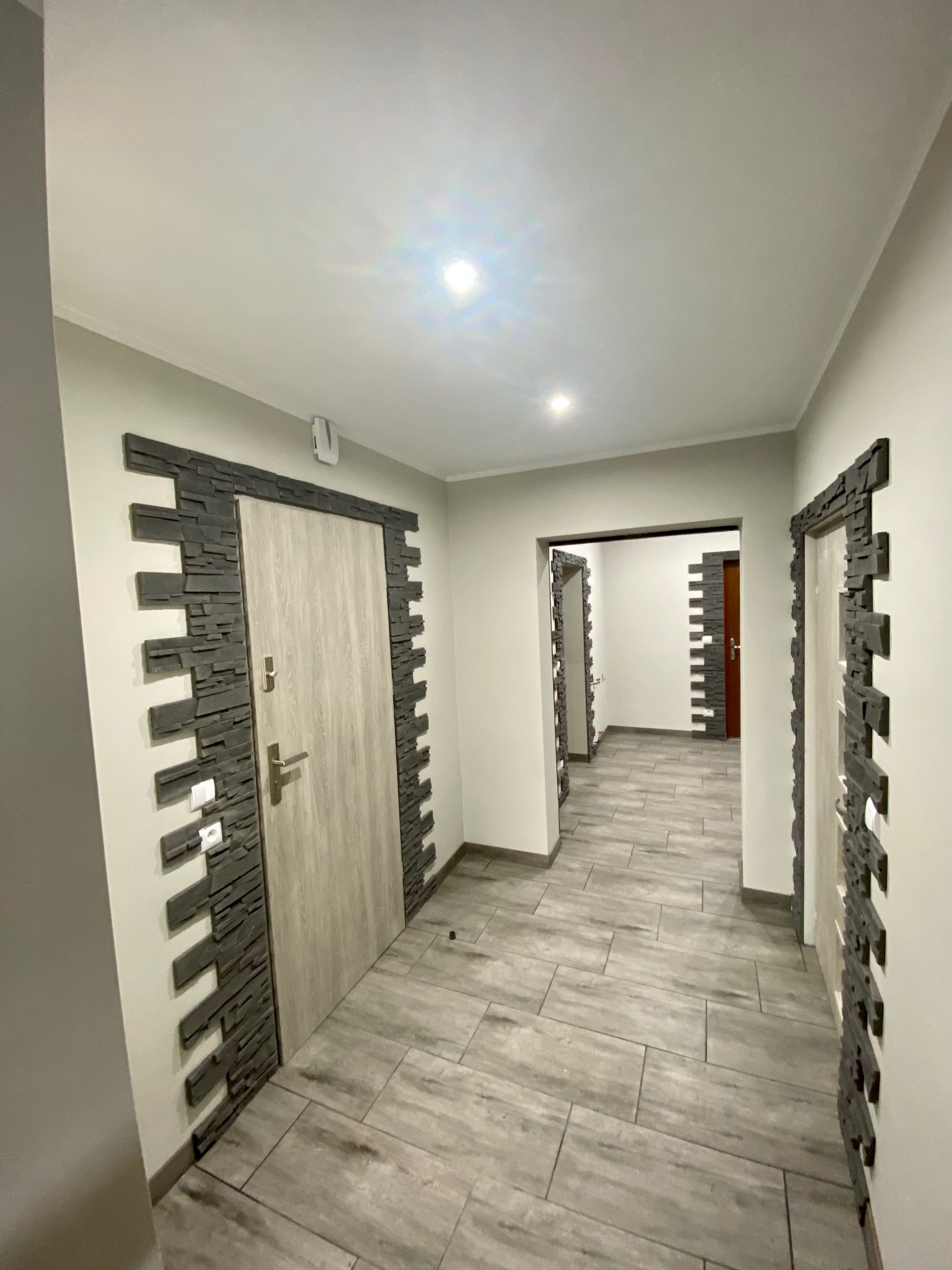 Wykończony korytarz z szarą podłogą imitującą drewno, drzwi obramowane dekoracyjną okładziną z szarego kamienia, widok perspektywiczny w głąb korytarza.