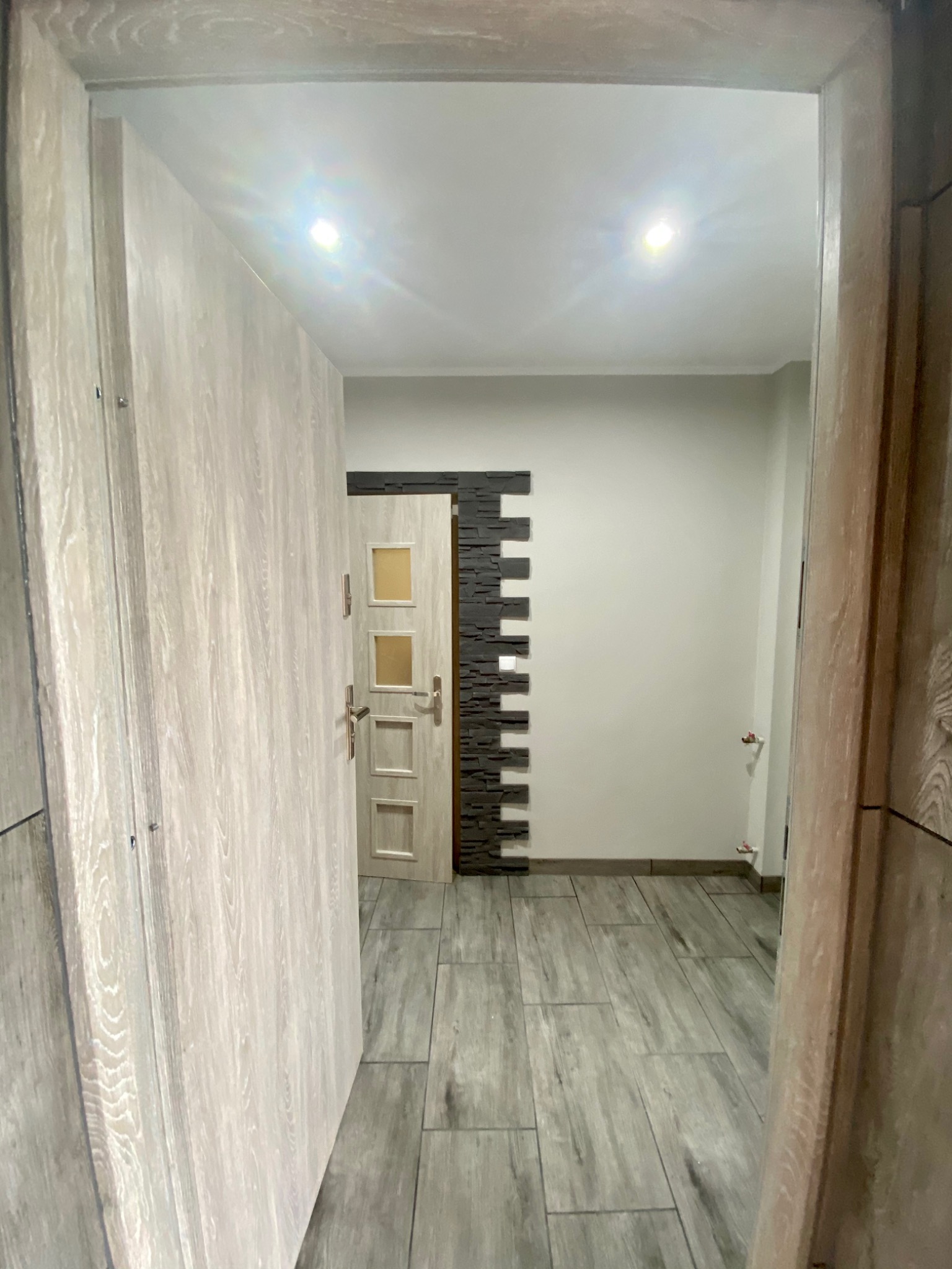 Wnętrze łazienki z szarą podłogą imitującą drewno, białe ściany z dekoracyjnym panelem z ciemnego kamienia, widok przez otwarte drzwi z okładziną imitującą drewno.