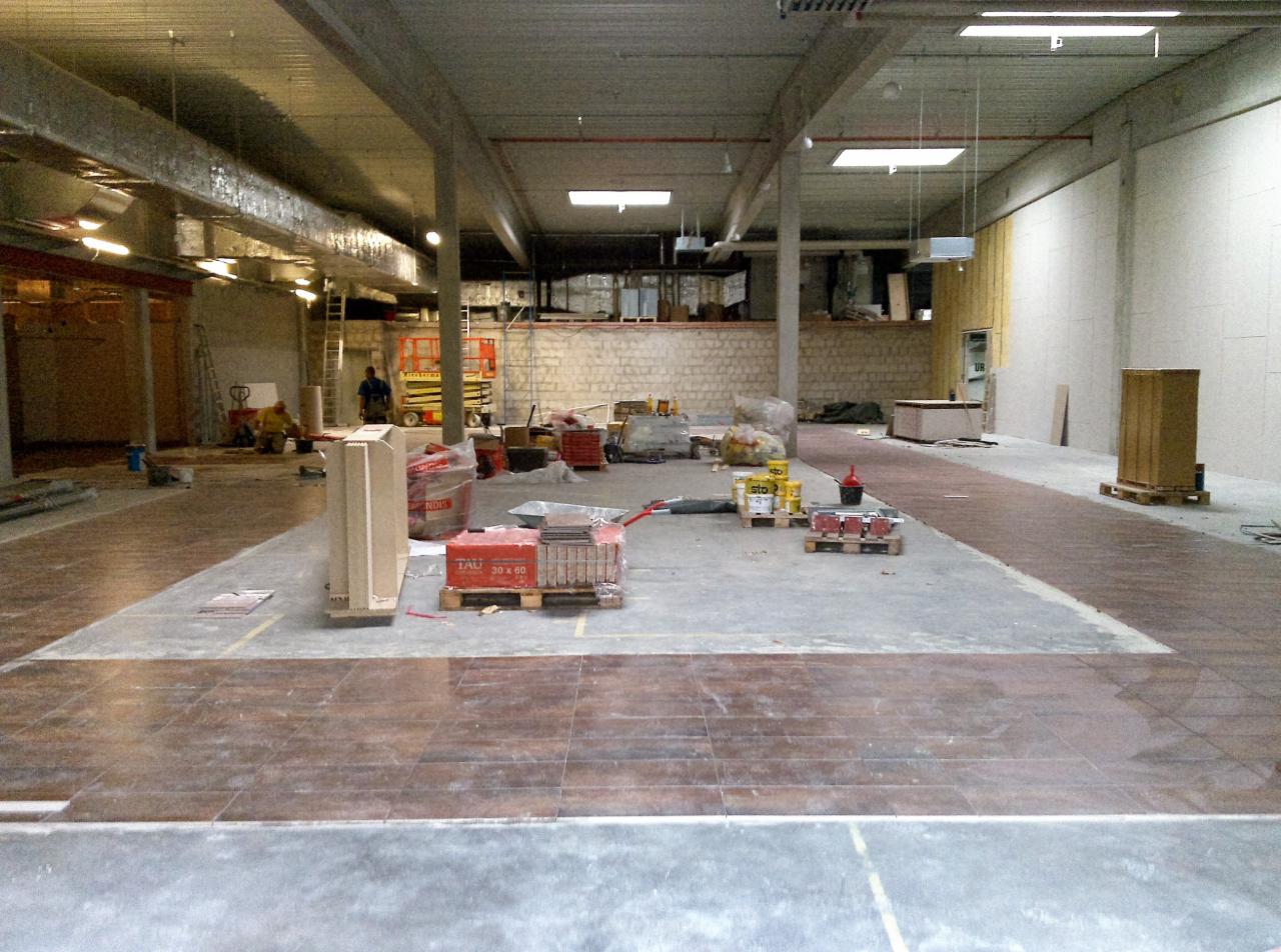 Rozległa przestrzeń komercyjna w trakcie układania brązowych płytek podłogowych, widoczne materiały budowlane, narzędzia i pracownicy w tle.