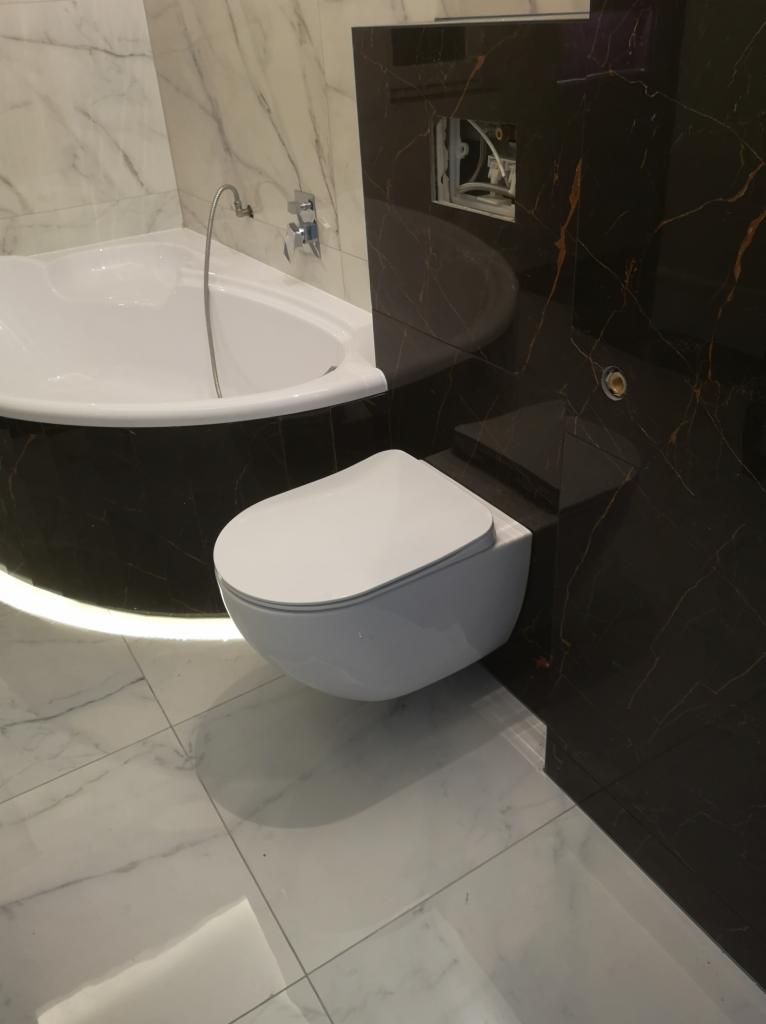 Nowoczesna łazienka z wiszącą toaletą, marmurową wanną z podświetleniem LED i czarnymi płytkami imitującymi marmur z złotymi żyłami.