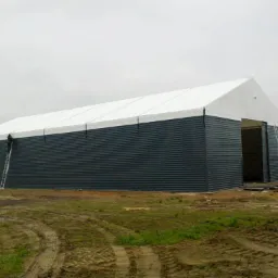 Hala z płyt warstwowych z białym dachem, drabina oparta o ścianę, widoczny pracownik. Teren wokół hali z błotem i śladami opon. Pochmurne niebo w tle.