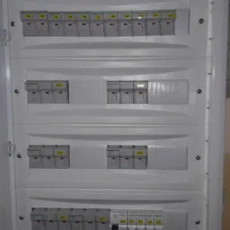 Otwarta, biała skrzynka rozdzielcza z zainstalowanymi bezpiecznikami i modułami elektronicznymi, widoczne oznaczenia Q1-Q18 oraz P1-P6.