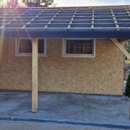 FHU A&W Agnieszka Wargocka - Konstrukcja budynku w trakcie budowy: szkielet dachu z łatami, ściany z płyt OSB, widoczne okna i słupy nośne, na pierwszym planie betonowa wylewka, w tle zieleń ogrodu.