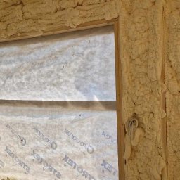 FHU A&W Agnieszka Wargocka - Aplikacja pianki poliuretanowej w nowym budynku, widoczne drewniane ramy okienne i ściany pokryte grubą warstwą izolacji natryskowej, z fragmentami kabli elektrycznych wystających spod piany.