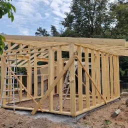 Konstrukcja drewniana domu w stanie surowym otwartym, z widocznymi elementami więźby dachowej i ścian szkieletowych, na tle zieleni w Osiecku. Na pierwszym planie ziemia i drabina.