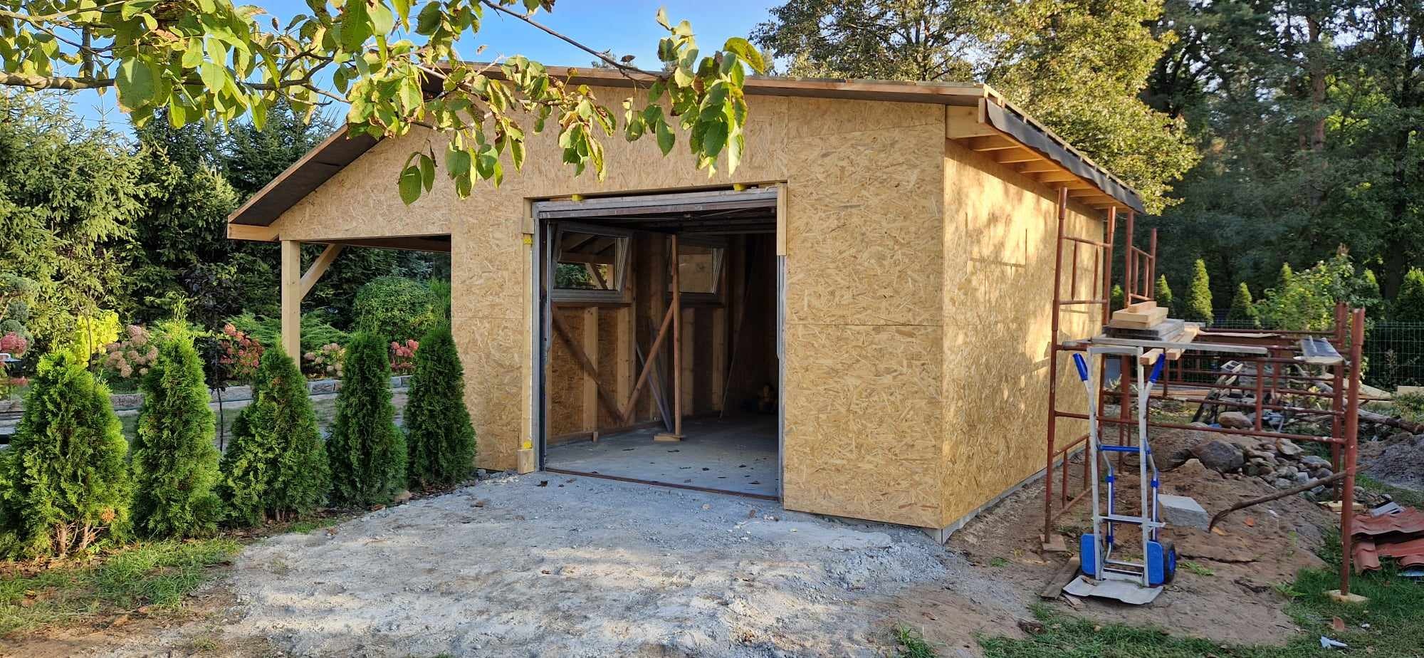 Surowy garaż z płyty OSB w trakcie budowy, z otwartymi wrotami, widoczne rusztowanie i wózek transportowy, zieleń w tle.