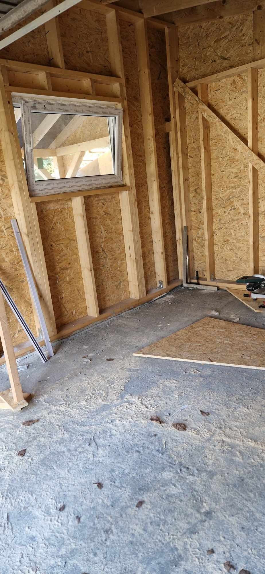 Wnętrze domu w trakcie budowy, widoczna konstrukcja szkieletowa z drewna i płyty OSB, okno z białymi ramami, narzędzia i fragmenty płyty na podłodze.