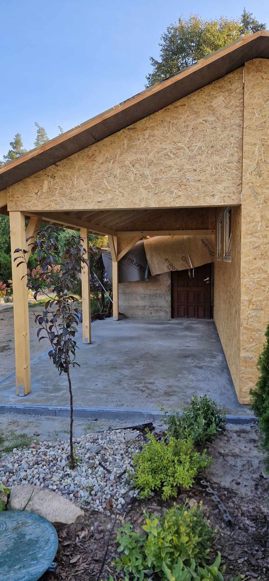 Surowy stan konstrukcji budynku z płyty OSB z widocznym zadaszeniem, betonową podłogą i młodym drzewkiem na pierwszym planie.