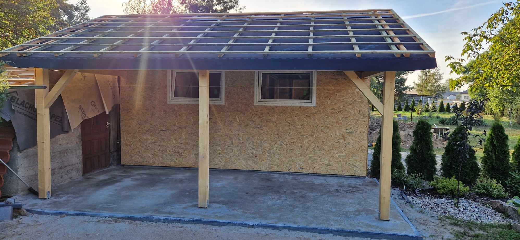 Konstrukcja budynku w trakcie budowy: szkielet dachu z łatami, ściany z płyt OSB, widoczne okna i słupy nośne, na pierwszym planie betonowa wylewka, w tle zieleń ogrodu.