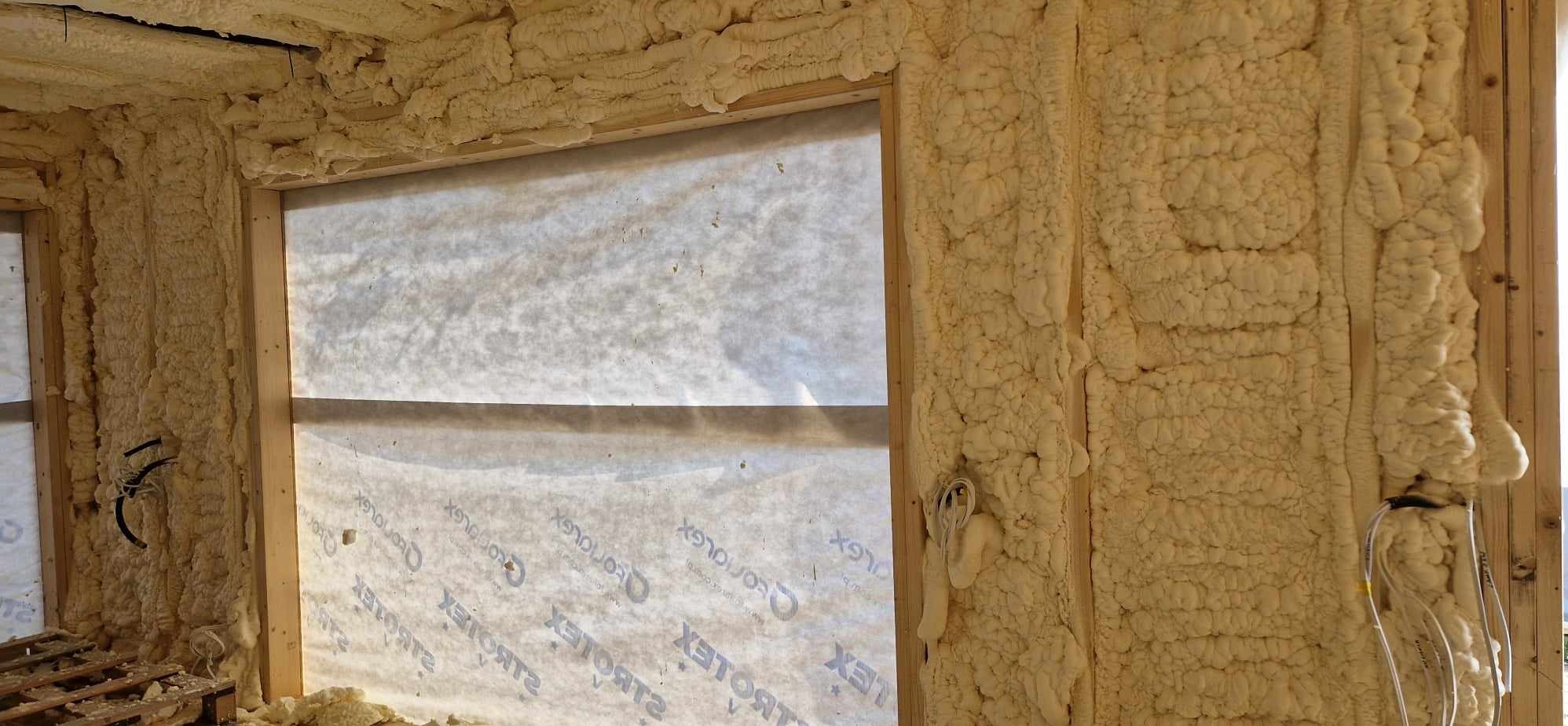 Aplikacja pianki poliuretanowej w nowym budynku, widoczne drewniane ramy okienne i ściany pokryte grubą warstwą izolacji natryskowej, z fragmentami kabli elektrycznych wystających spod piany.
