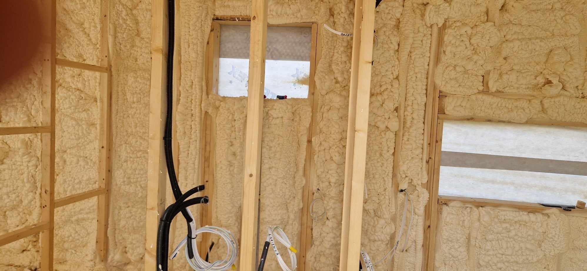 Wnętrze domu w trakcie budowy, widoczne drewniane ramy ścian wypełnione pianą izolacyjną, okno zabezpieczone folią, instalacja elektryczna w peszlach i przewody.