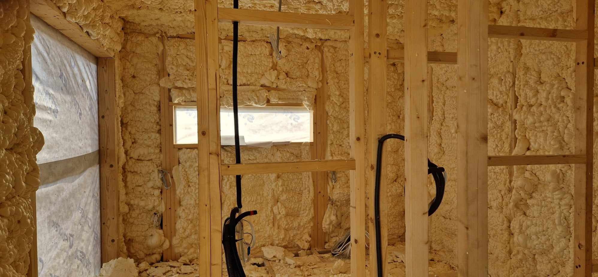 Wnętrze domu szkieletowego w trakcie ocieplania pianą PUR, widoczne drewniane belki konstrukcyjne, instalacje elektryczne w peszlach oraz okno z folią ochronną.