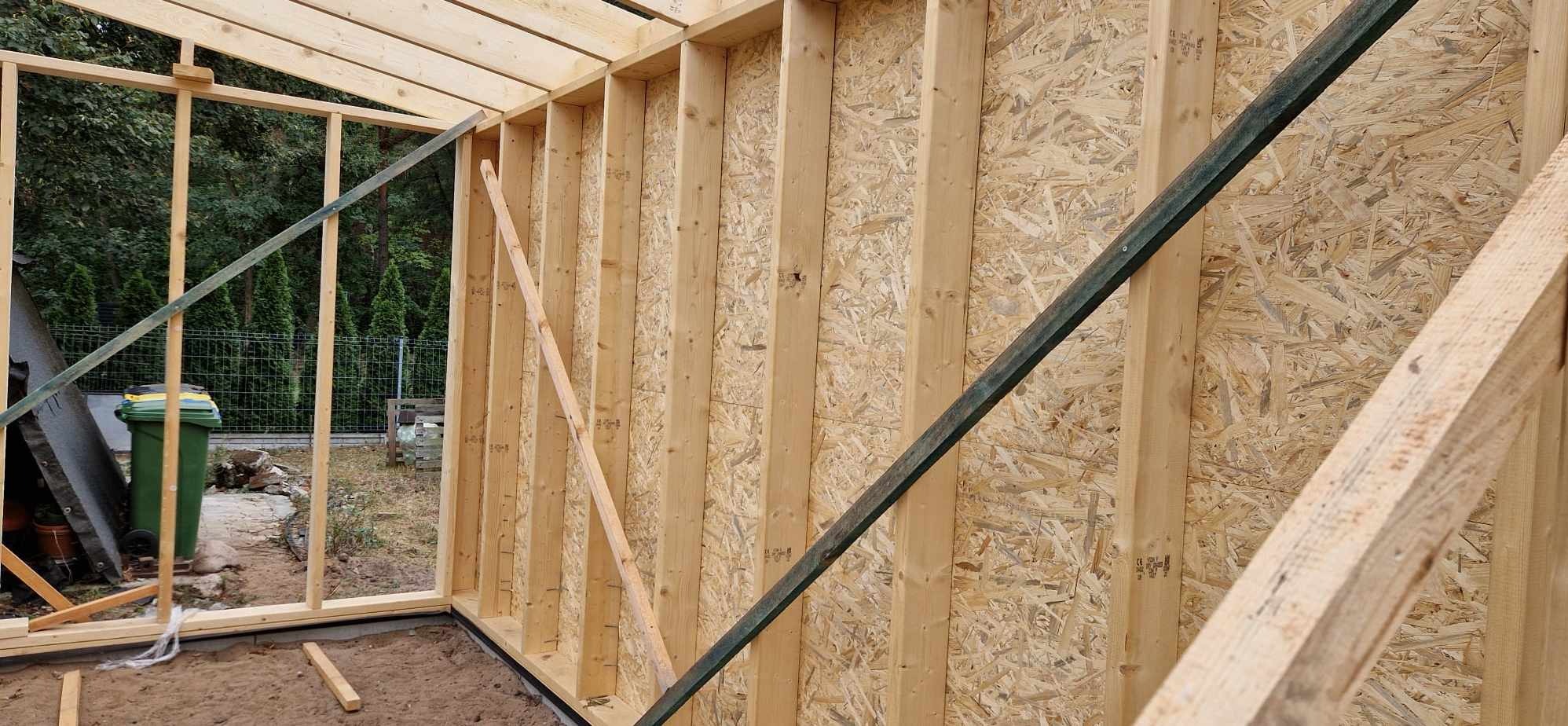 Wnętrze konstrukcji w trakcie budowy, widoczne drewniane szkielety ścian i płyty OSB, na zewnątrz zieleń i śmietnik.