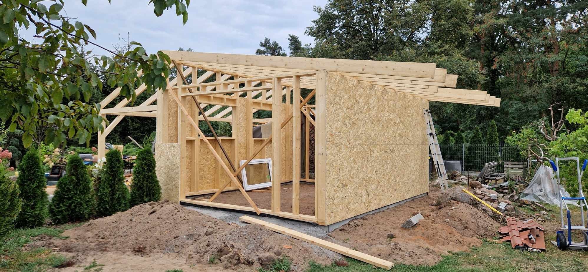 Konstrukcja szkieletowa budynku w trakcie budowy, z widocznymi elementami drewnianymi, płytami OSB na ścianach, oknem w ramie, oraz stertą ziemi i zielenią w tle.