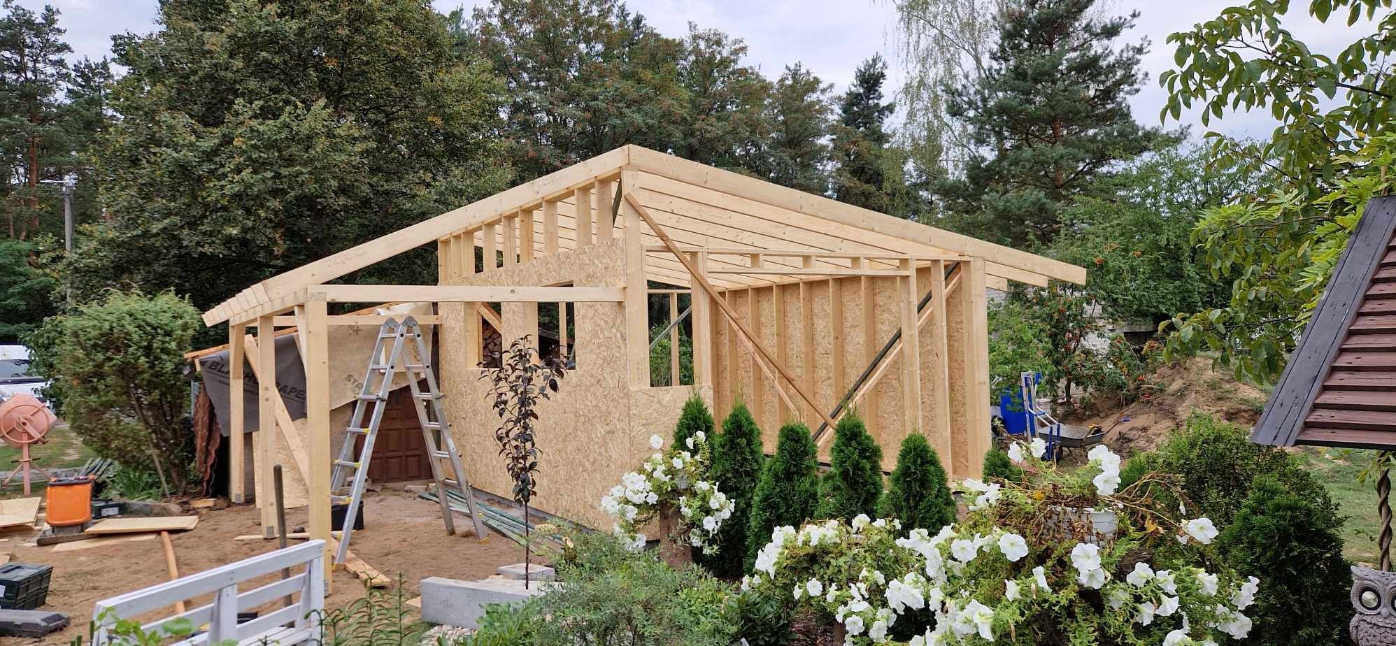 Konstrukcja domu w trakcie budowy, widoczne drewniane elementy szkieletu i płyty OSB, drabina oparta o ścianę, w tle zieleń drzew i krzewów.