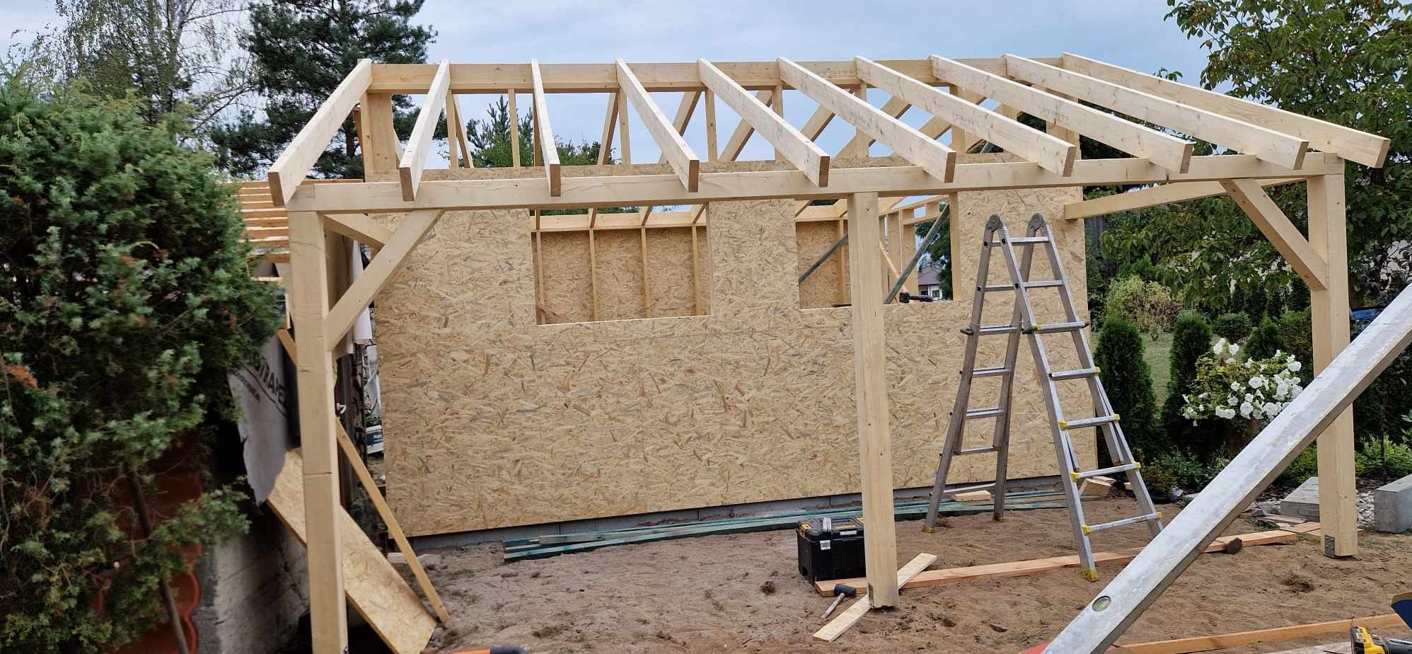 Wznoszenie konstrukcji szkieletowej budynku z drewnianych belek i płyt OSB, widoczne elementy dachu i ściany z otworami okiennymi, drabina oparta o ścianę, narzędzia na ziemi.