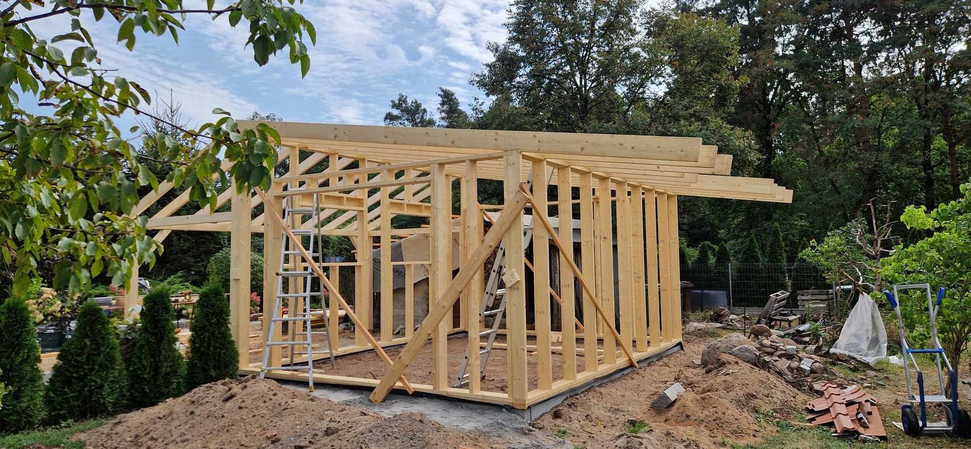 Konstrukcja drewniana domu w stanie surowym otwartym, z widocznymi elementami więźby dachowej i ścian szkieletowych, na tle zieleni w Osiecku. Na pierwszym planie ziemia i drabina.