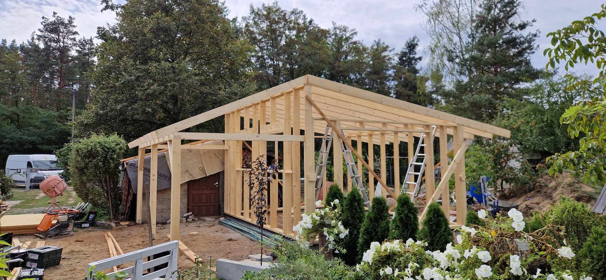 Szkielet drewnianej konstrukcji w ogrodzie, widoczne deski, drabiny, materiały izolacyjne i roślinność, w tle las i samochód dostawczy.