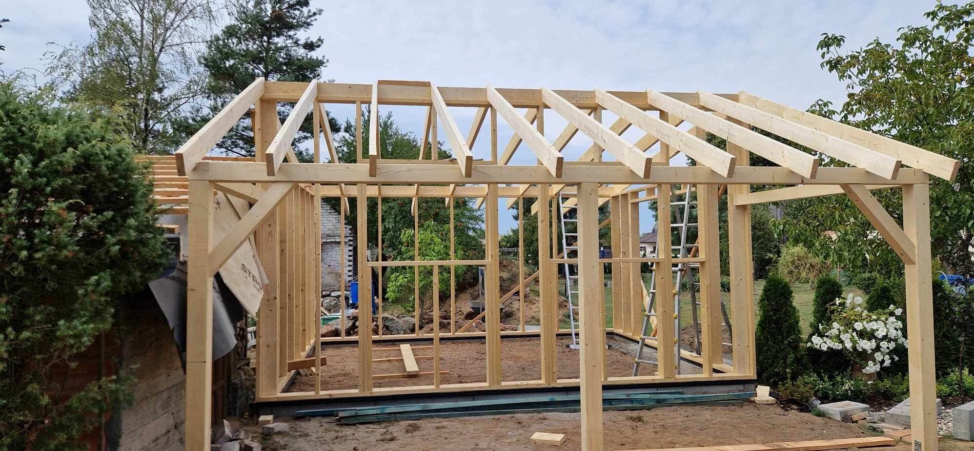 Szkieletowa konstrukcja drewniana w trakcie budowy, widoczne belki stropowe i ściany, drabina oparta o jedną ze ścian, w tle zieleń drzew i krzewów.