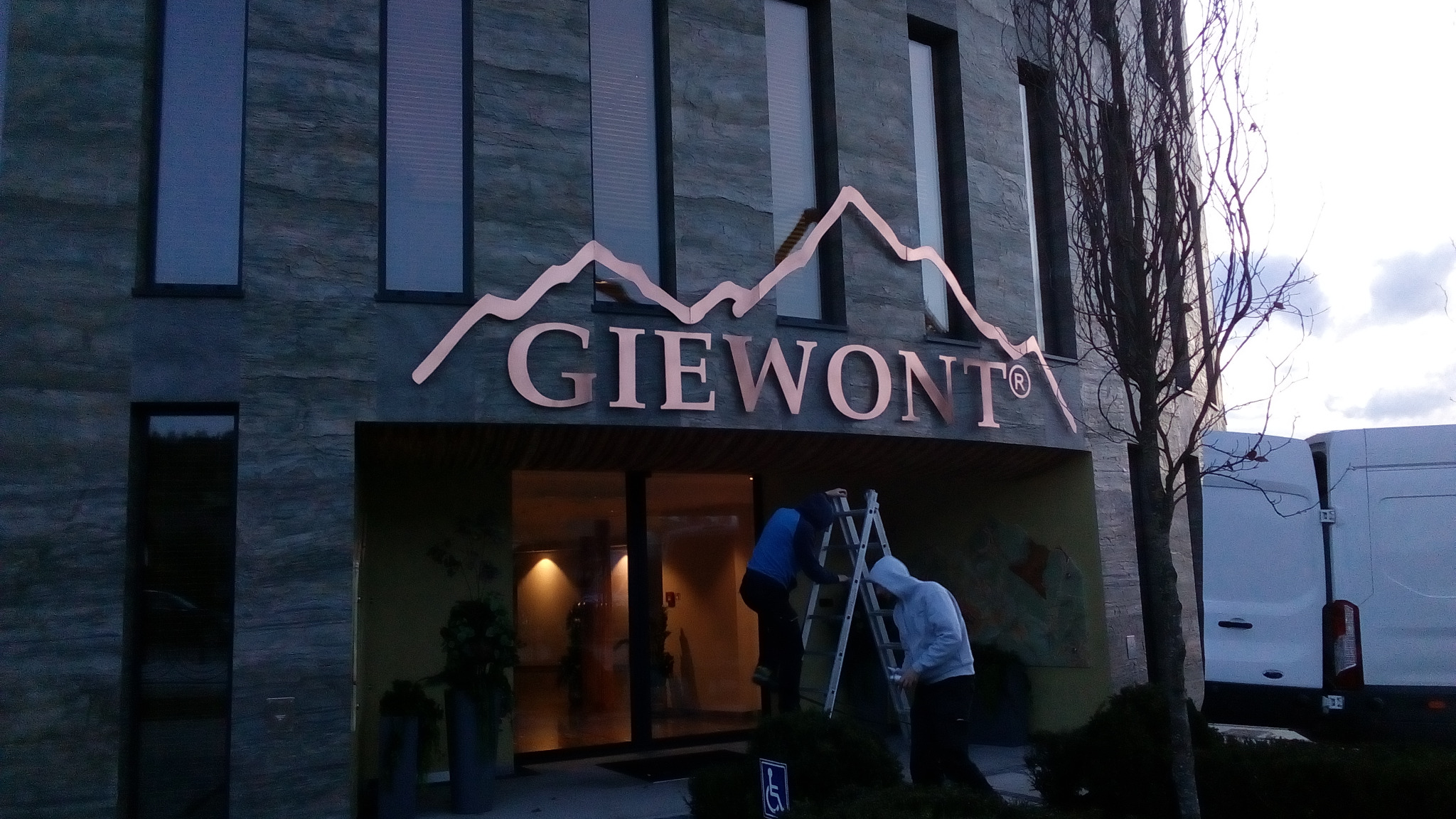 Montaż miedzianego szyldu z nazwą 'GIEWONT' i symbolem górskim nad wejściem do budynku, pracownicy na drabinie, biały bus zaparkowany obok.
