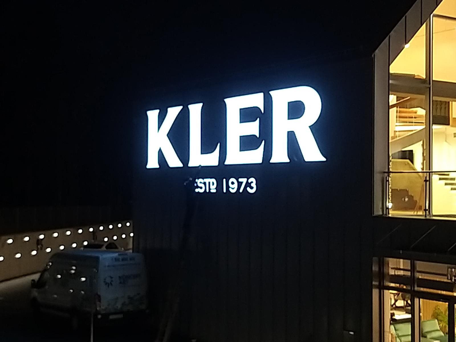 Podświetlany szyld z nazwą 'KLER ESTD 1973' na ciemnej fasadzie budynku, obok zaparkowany samochód dostawczy.
