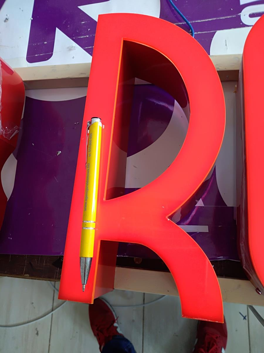 Czerwona litera 'R' z tworzywa sztucznego, podświetlana od wewnątrz, oparta o żółty długopis dla porównania skali, na tle fioletowego baneru z białymi elementami graficznymi.