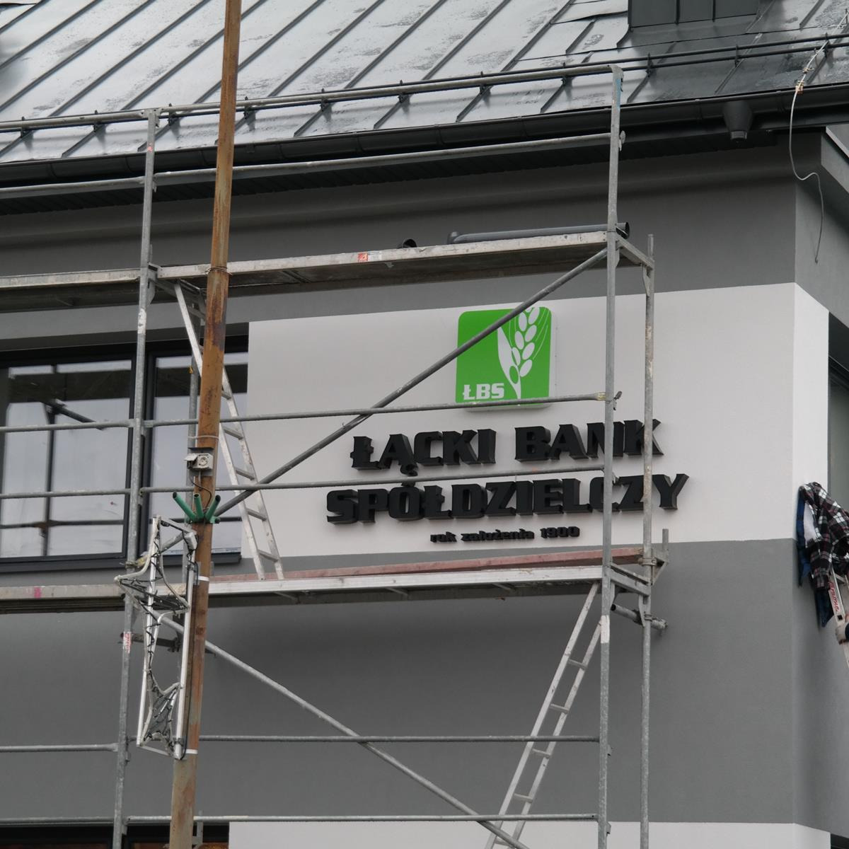 Montaż szyldu Łącki Bank Spółdzielczy na elewacji budynku, widoczne rusztowanie.