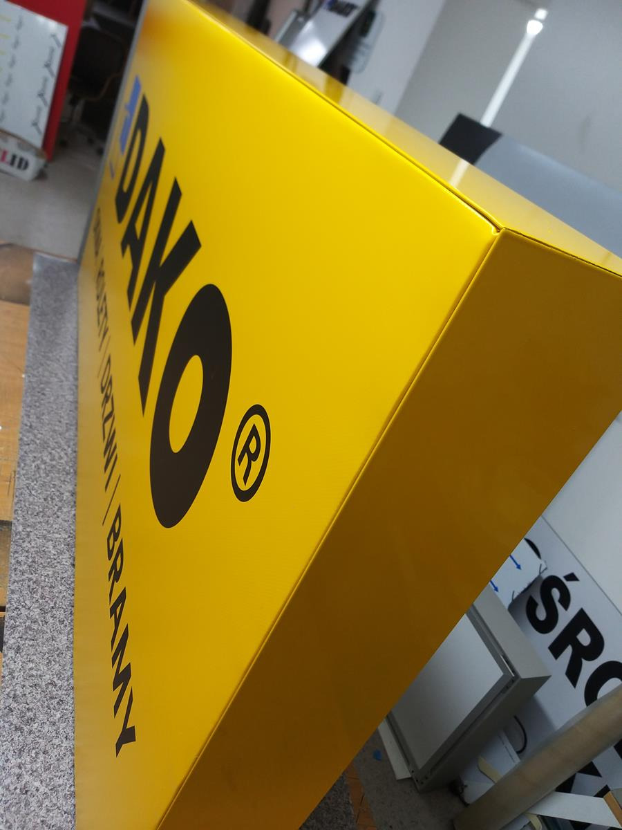 Żółty kaseton reklamowy z czarnym napisem 'DAKO' i logo, widziany pod kątem w warsztacie.