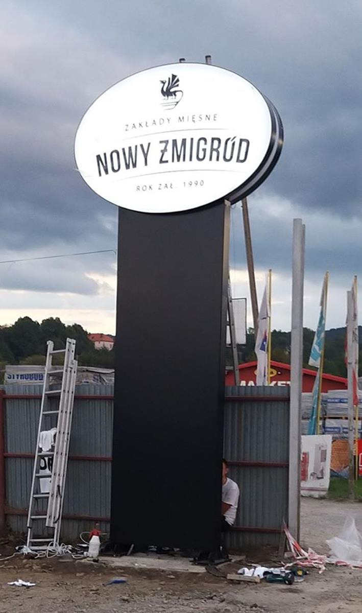 Montaż podświetlanego pylonu reklamowego z owalnym szyldem 'Zakłady Mięsne Nowy Żmigród' na tle pochmurnego nieba, drabina oparta o ogrodzenie, widoczny fragment osoby podczas instalacji.