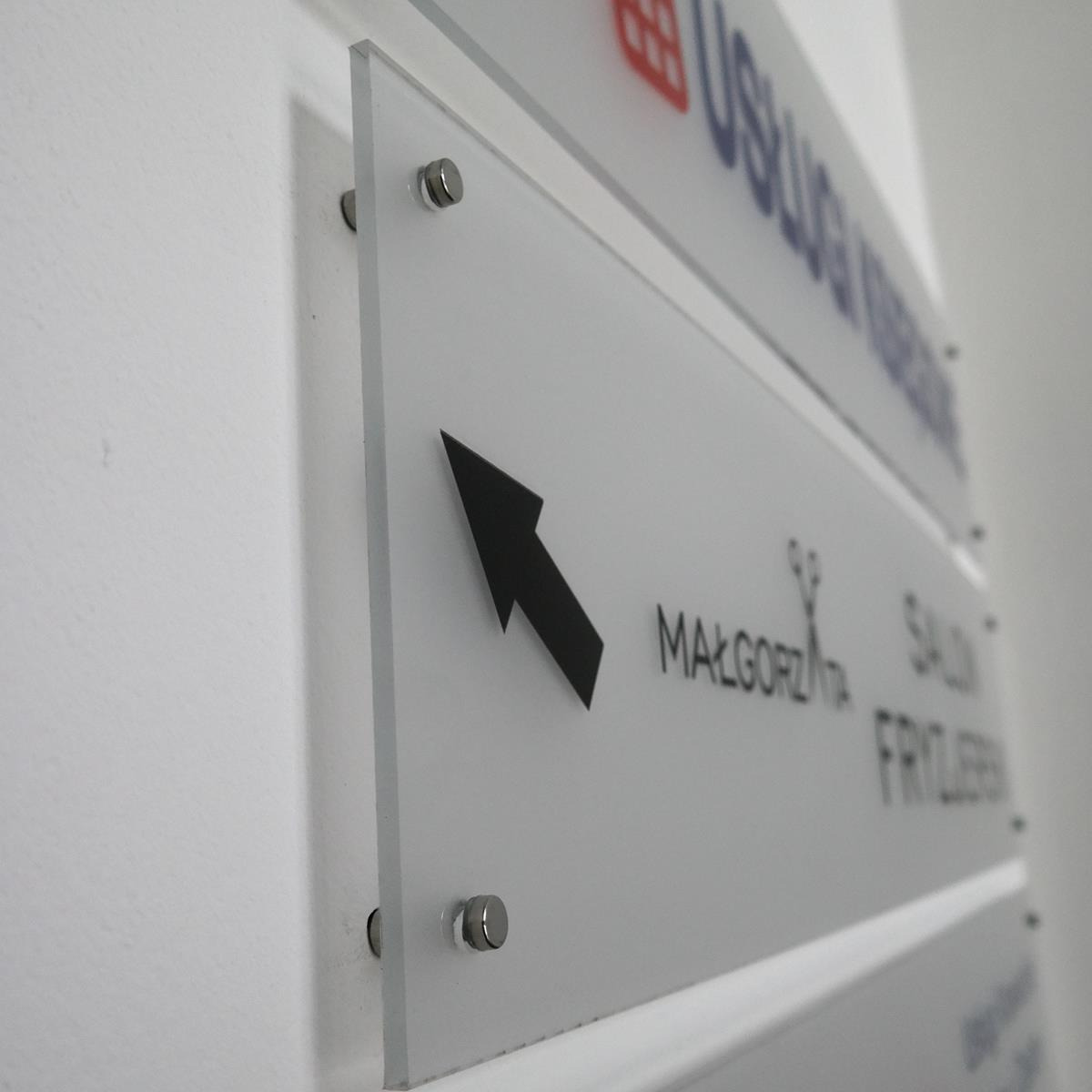 Szklany szyld informacyjny z czarną strzałką wskazującą kierunek do salonu fryzjerskiego Małgorzata, zamocowany na ścianie za pomocą metalowych śrub dystansowych.