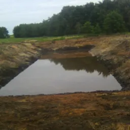 Świeżo wykopany, prostokątny staw z wodą, otoczony brzegiem z ziemi, w tle widoczny las i pochmurne niebo.