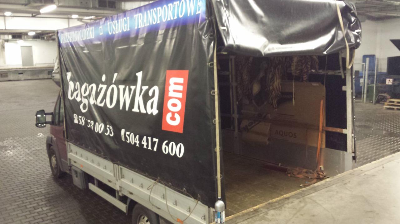 Ciężarówka z otwartą przestrzenią ładunkową, wypełniona kartonami i zabezpieczona plandeką z logo firmy transportowej bagażówka.com. Widoczny numer telefonu.