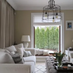 Jasny salon z kremowymi ścianami, białymi meblami i dużym oknem z widokiem na wysokie tuje, czarna metalowa lampa wisząca, szary puf z tacą i tulipanami.