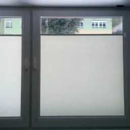 Zamknięte okno dwuskrzydłowe z zamontowanymi roletami zaciemniającymi w jasnym kolorze, widok z wnętrza pomieszczenia w Łodzi.